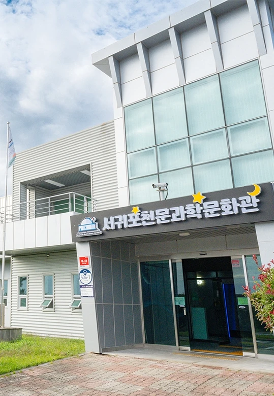 흐린 하늘 아래 회색빛 외관의 서귀포천문과학문화관 건물 입구와 달, 별 장식이 달린 간판이 보이는 외부 전경