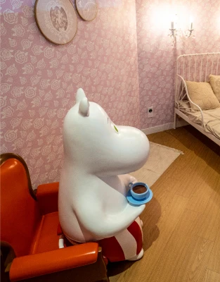 분홍 벽지로 꾸민 방에서 주황 의자에 앉아 찻잔을 든 무민 캐릭터 조형물