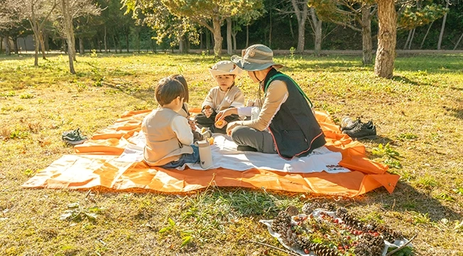 주황 돗자리에 둘러앉은 어른과 두 아이가 열매로 가을 피자를 만들며 즐기는 모습